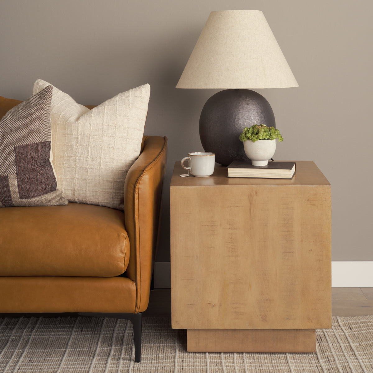 Brown leather sofa with decorative pillows and a wooden side table with lamp, mug, and plant.