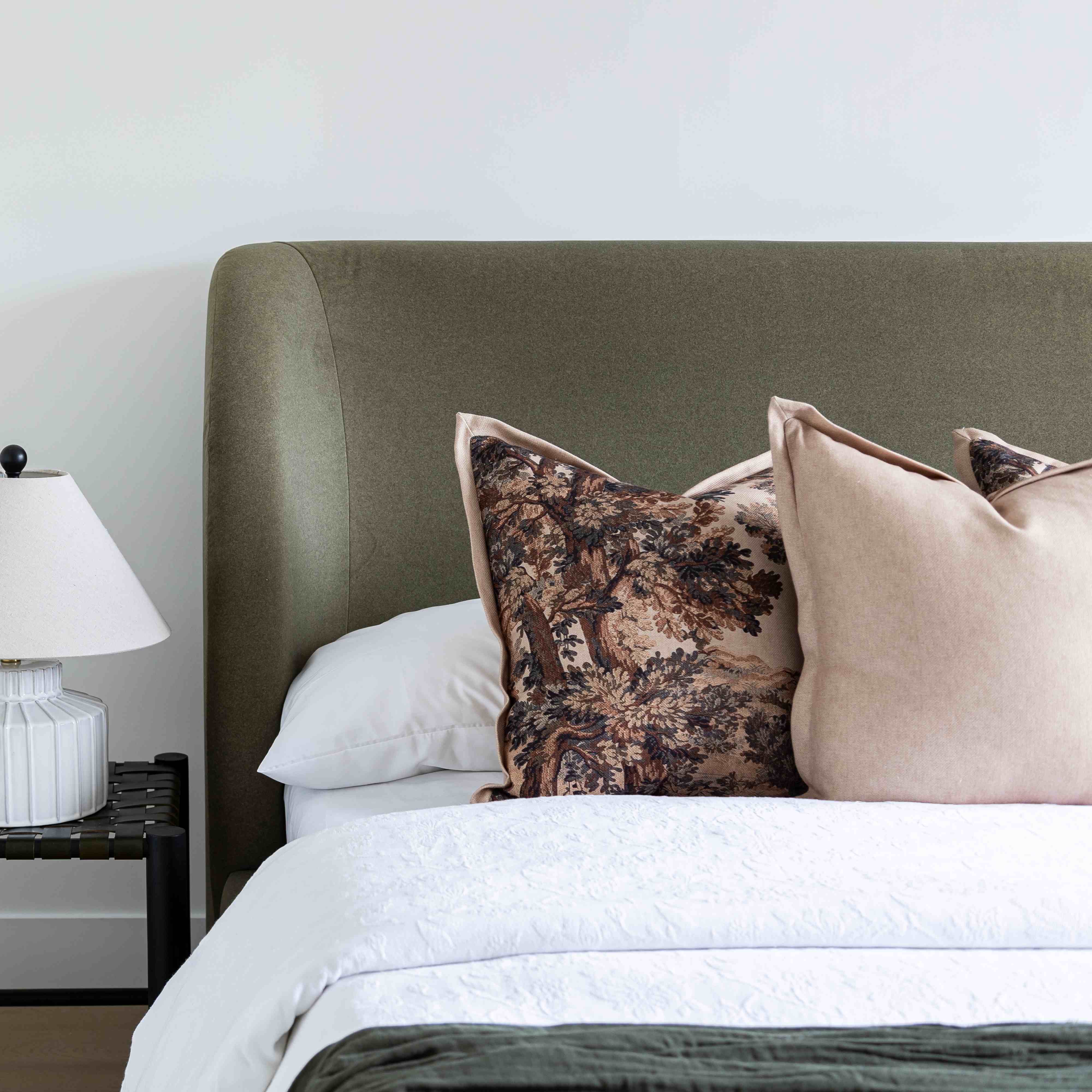 A styled bedroom scene showing the Aveline Velvet Bed with a tall upholstered headboard, white bedding, and two decorative pillows in front