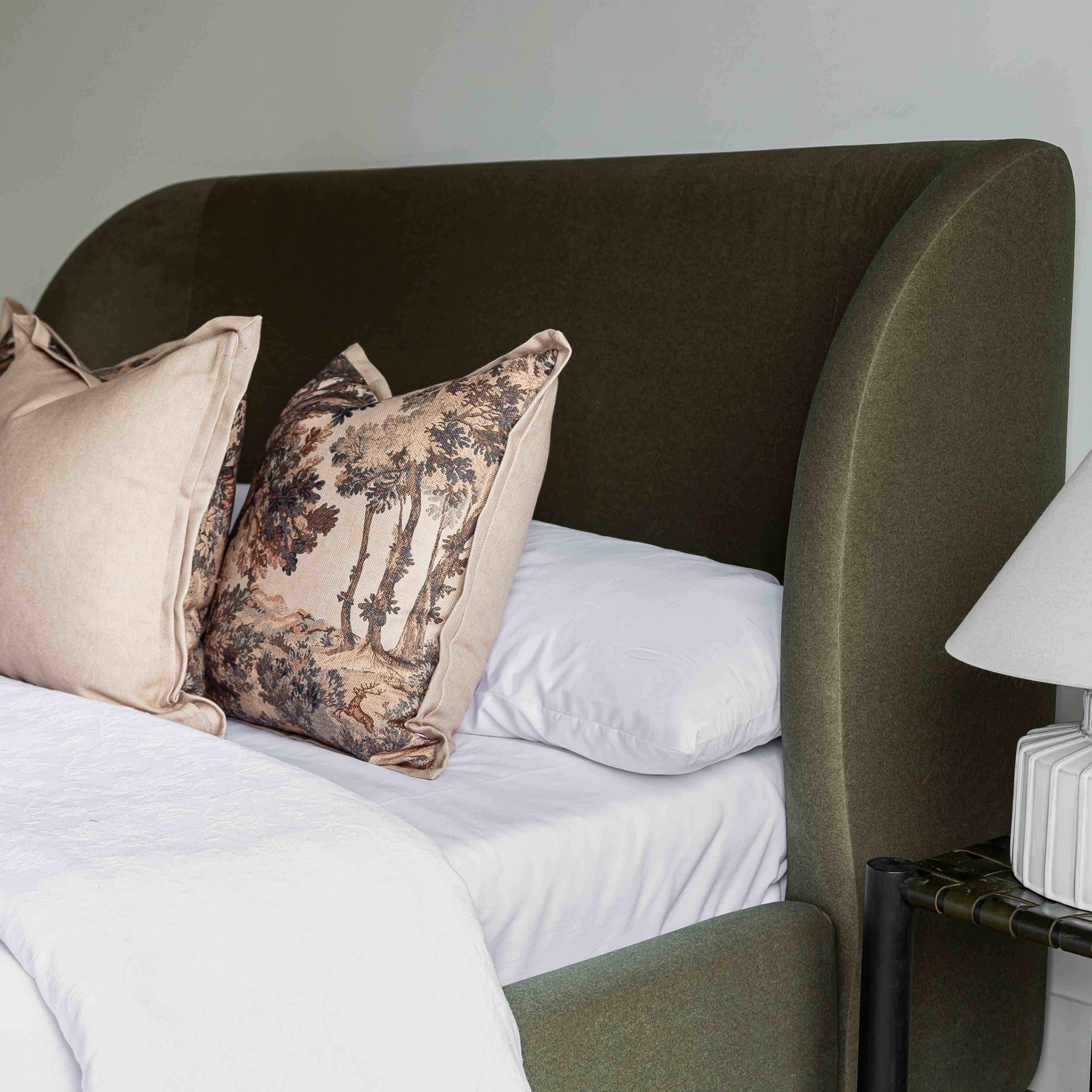 Aveline bed with a tall curved headboard in dark olive green, styled with white bedding, two beige pillows, and one patterned accent pillow, next to a bedside table and lamp.
