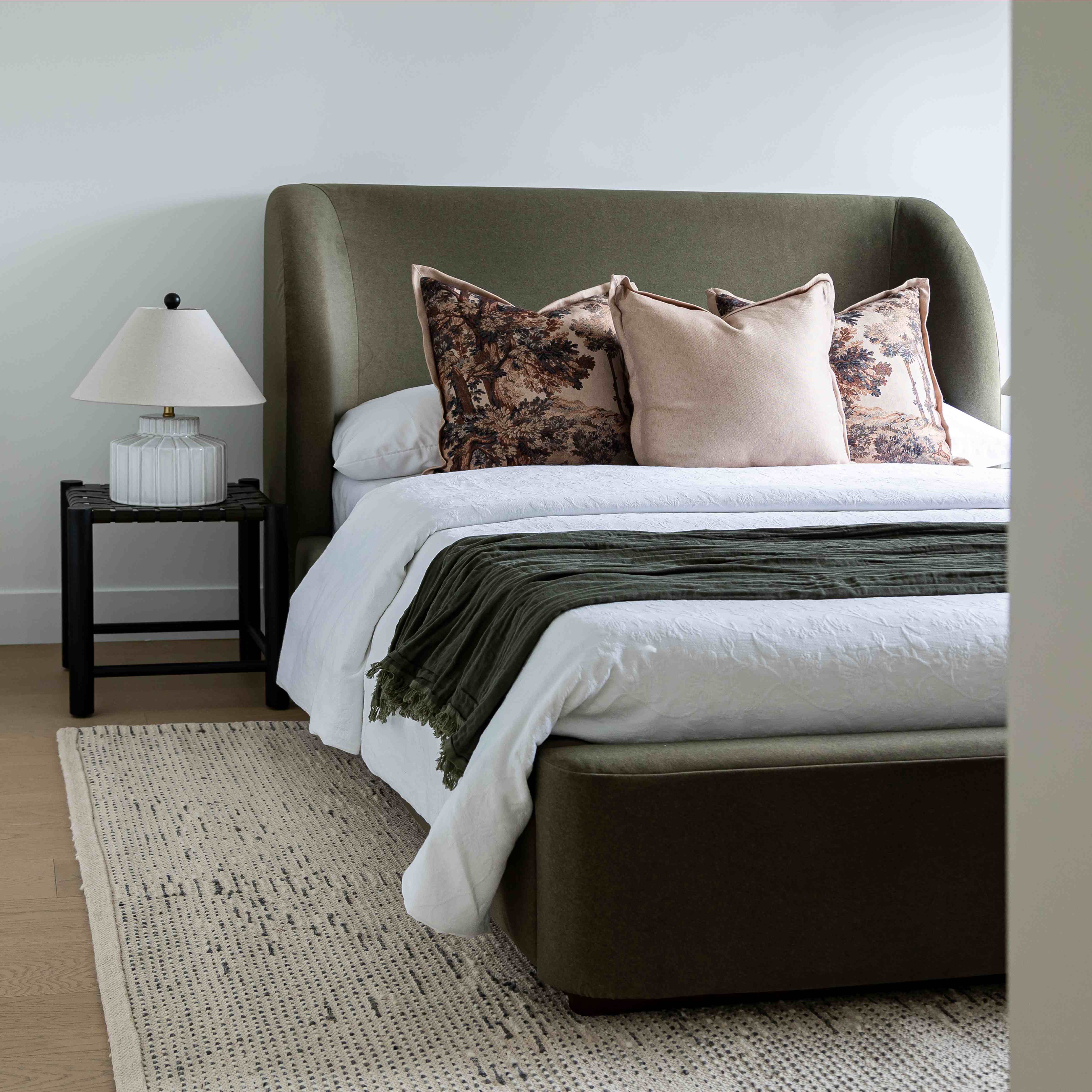 Aveline Velvet bed with a tall curved headboard and matching footboard in dark olive green, styled with white bedding, floral accent pillows, and a dark green throw blanket, set in a bedroom with a nightstand and lamp.