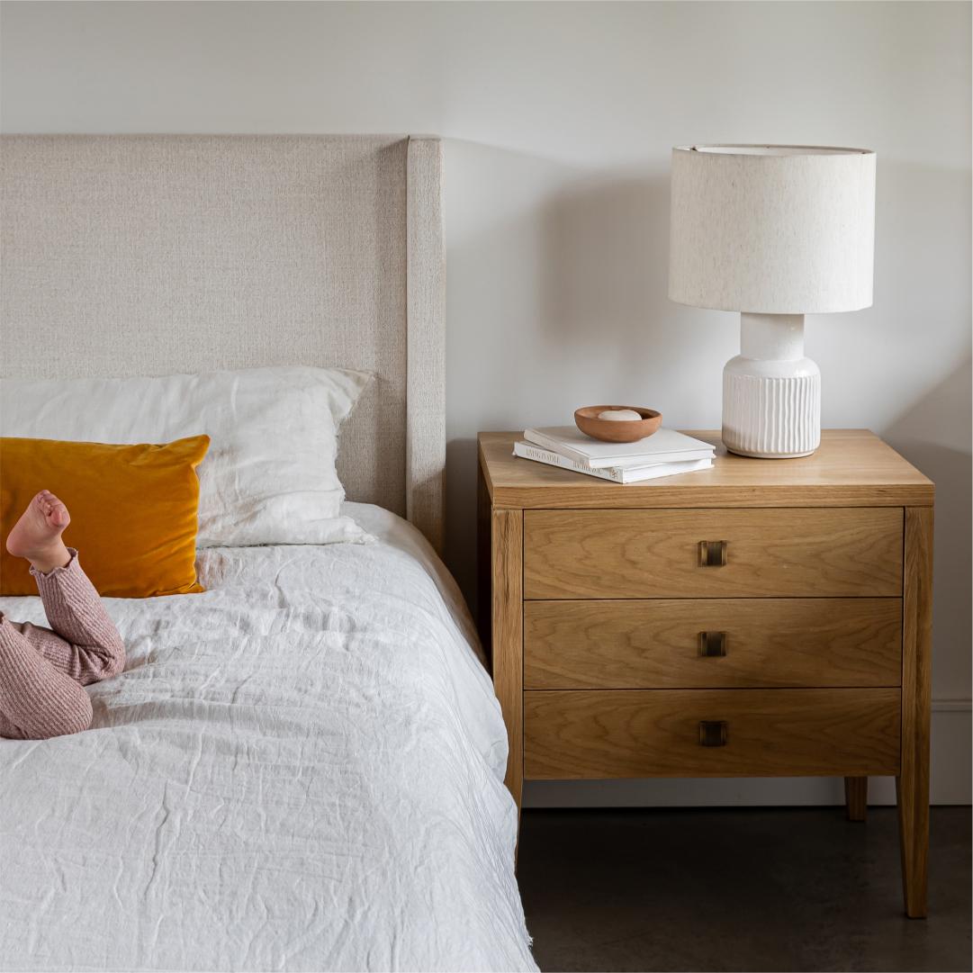 A close-up lifestyle shot of the Beaumont 3-Drawer Nightstand in Natural Oak positioned next to a beige upholstered bed. A white ceramic lamp and a small wooden bowl sit atop the nightstand.