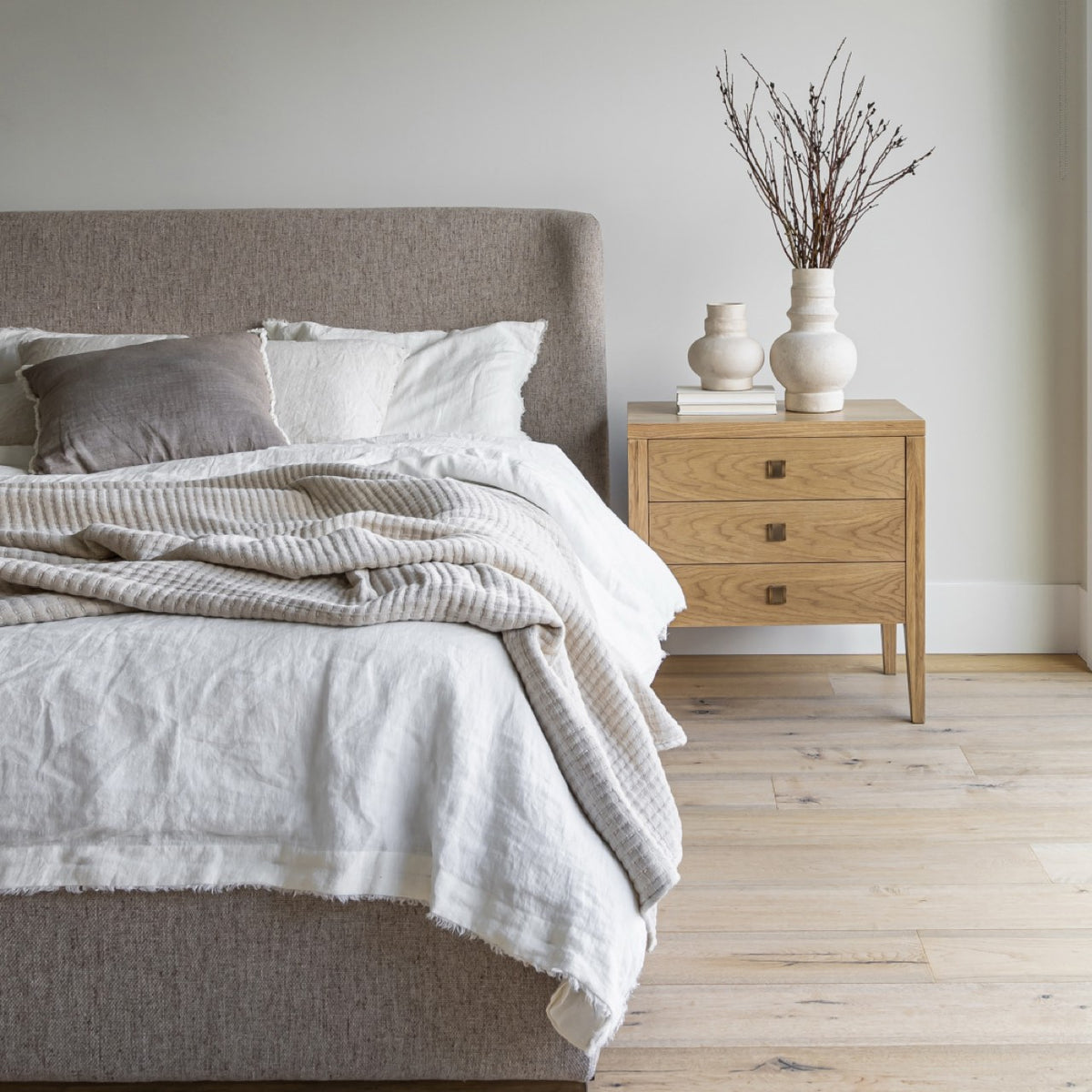 Wide view of the Beaumont 3-Drawer Nightstand in a modern bedroom with light oak flooring. The nightstand is styled with tall decorative branches in a textured vase.