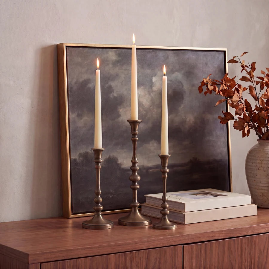 Caldria antique brass-finished candle holders styled with lit ivory taper candles on a wood console beside books and artwork.