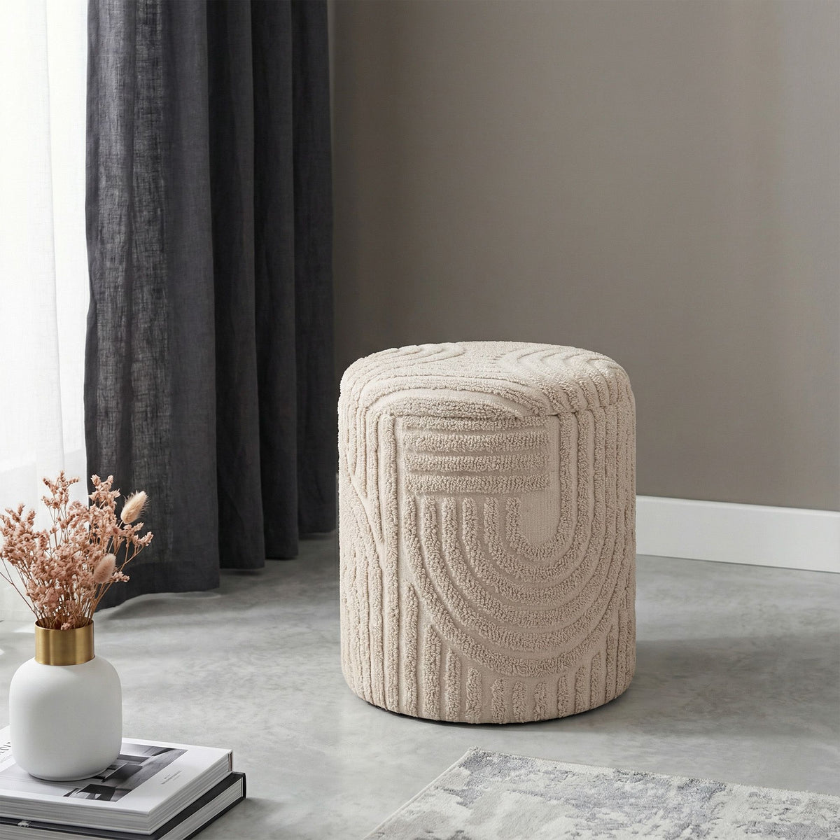 Beige textured ottoman in a room with a curtain and decor items.
