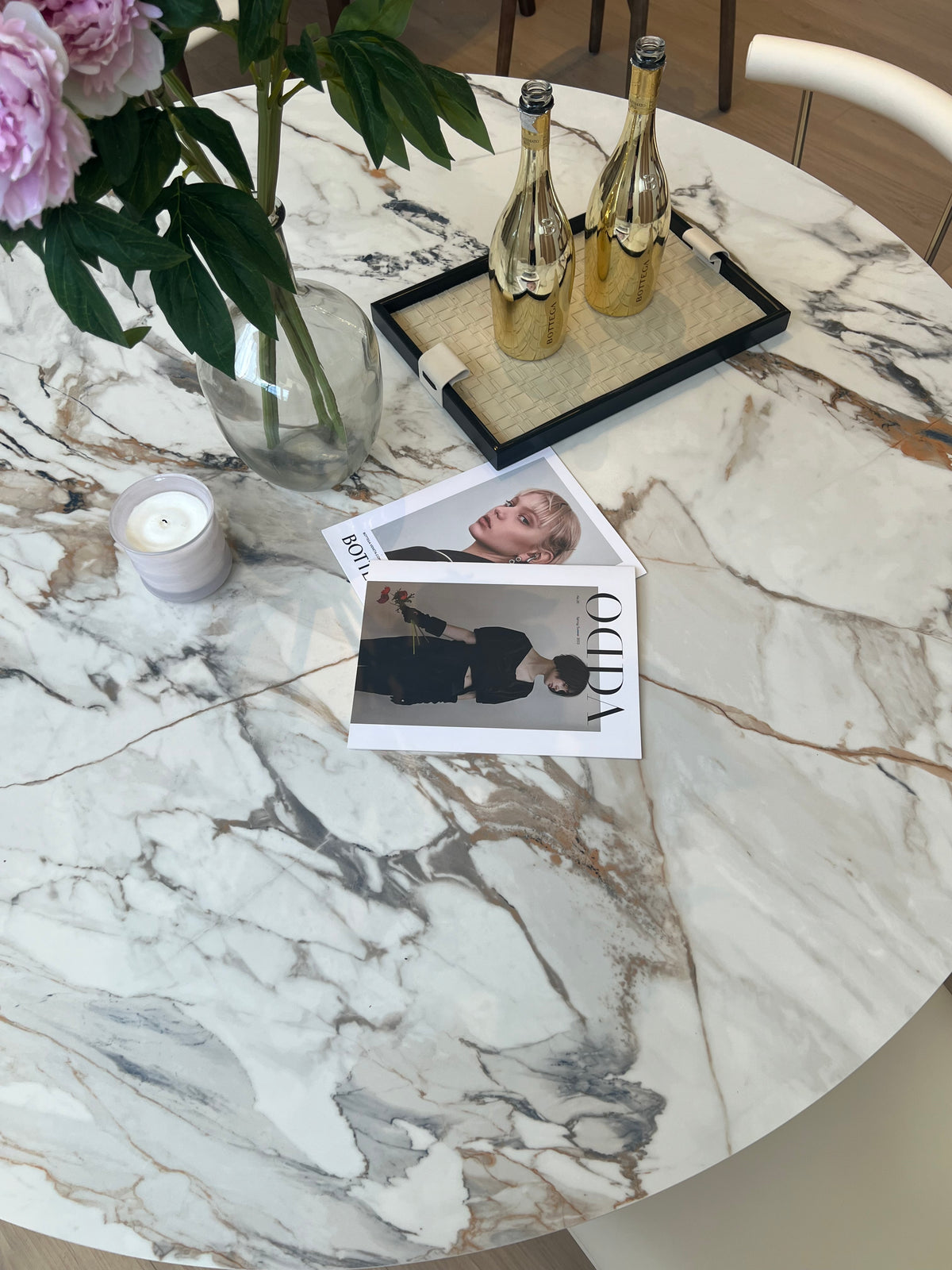 Close-up of Celestia Elegance round dining table with marble-patterned sintered stone top styled with flowers, magazines, and gold bottles