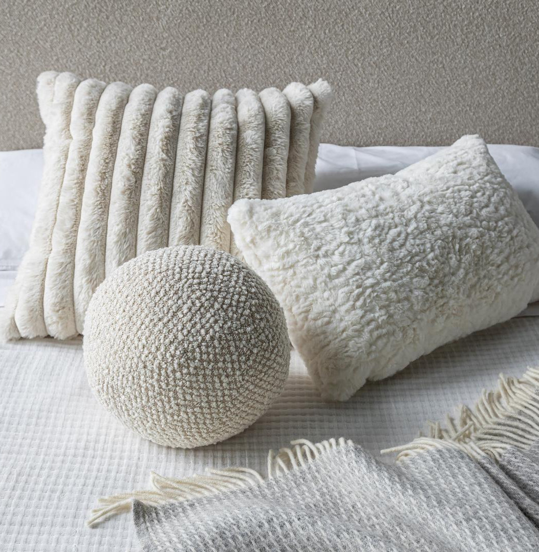A cozy bedroom setup featuring the Cloudline cushion trio layered on white bedding with a textured grey throw blanket.