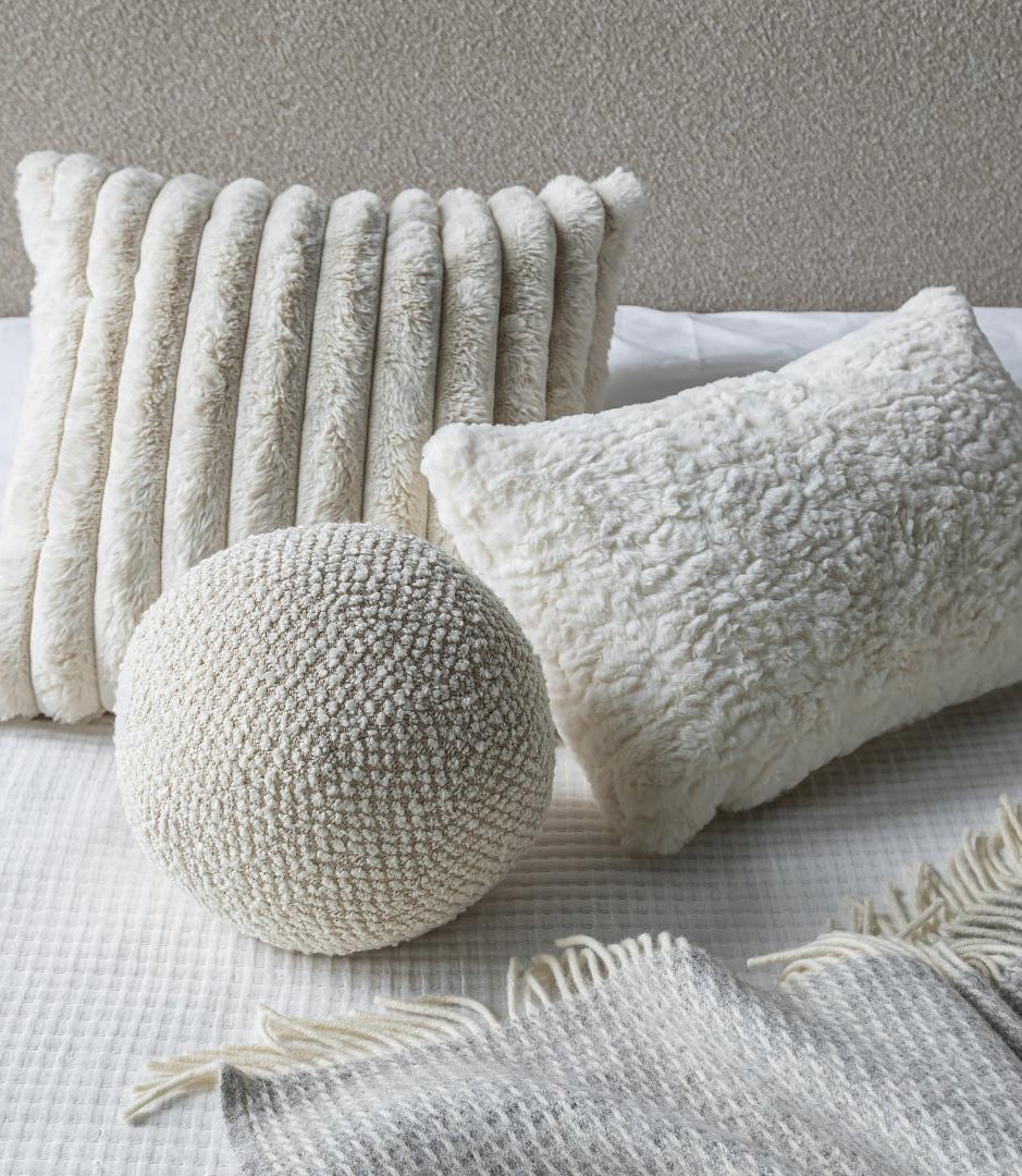 A cozy bedroom setup featuring the Cloudline cushion trio layered on white bedding with a textured grey throw blanket.