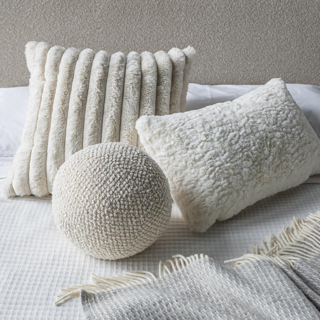 A cozy bedroom setup featuring the Cloudline cushion trio layered on white bedding with a textured grey throw blanket.