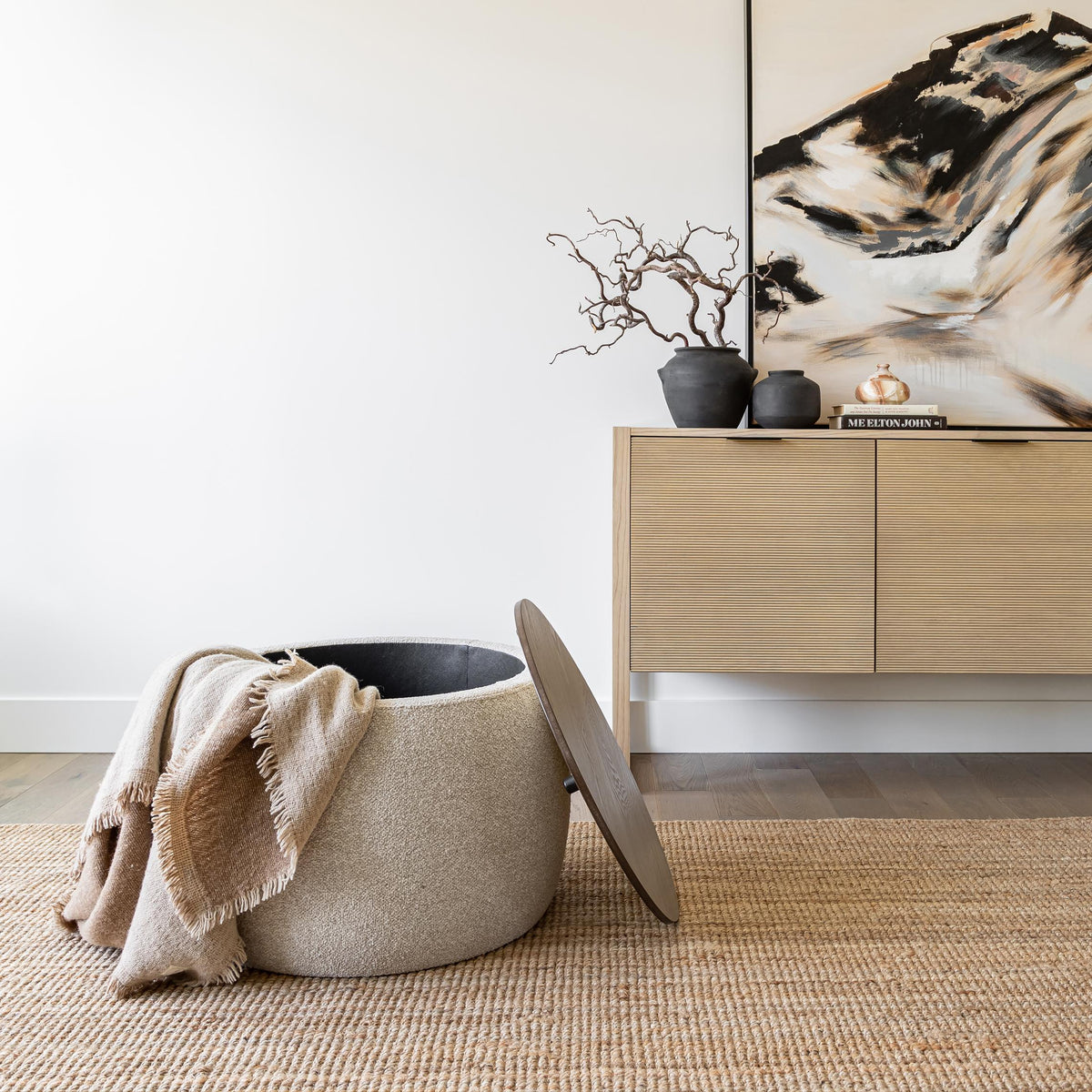 The Haven Storage Ottoman with the wood top removed to show blankets stored inside, styled in a bright minimalist room.