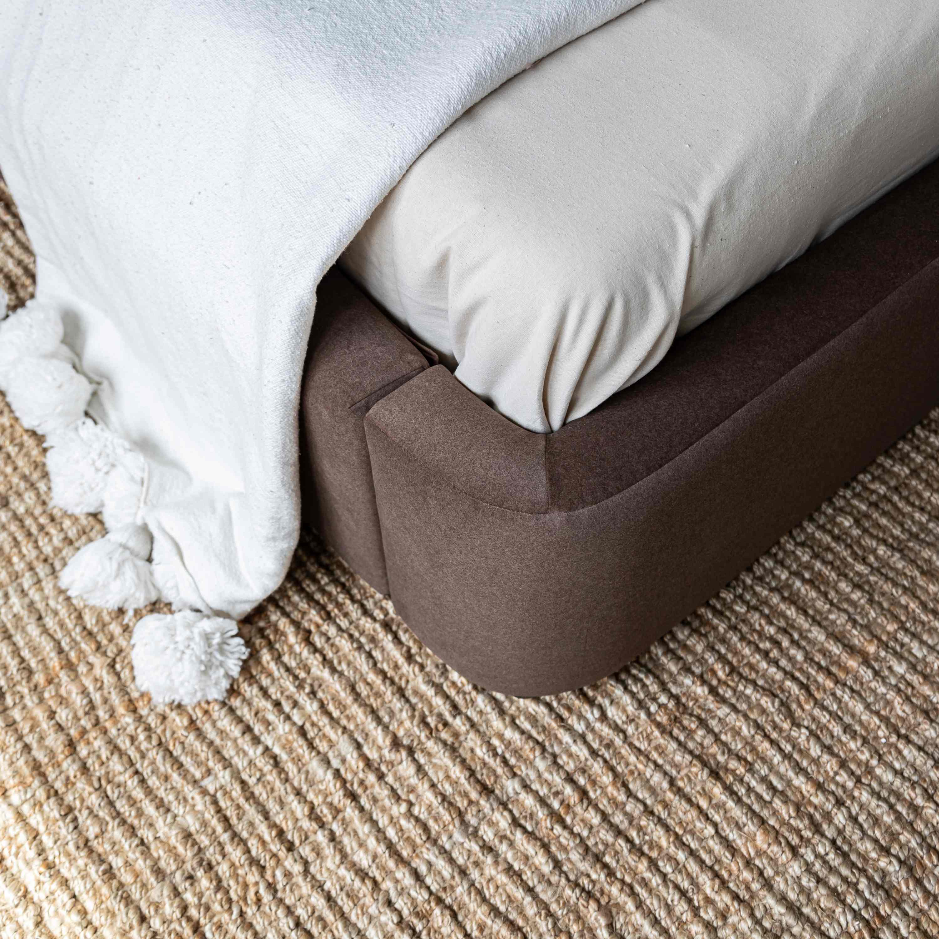 A close-up of the Lunelle bed corner with velvet upholstered bed frame, cream bedding, a white throw blanket with pom-poms, and a textured beige rug.