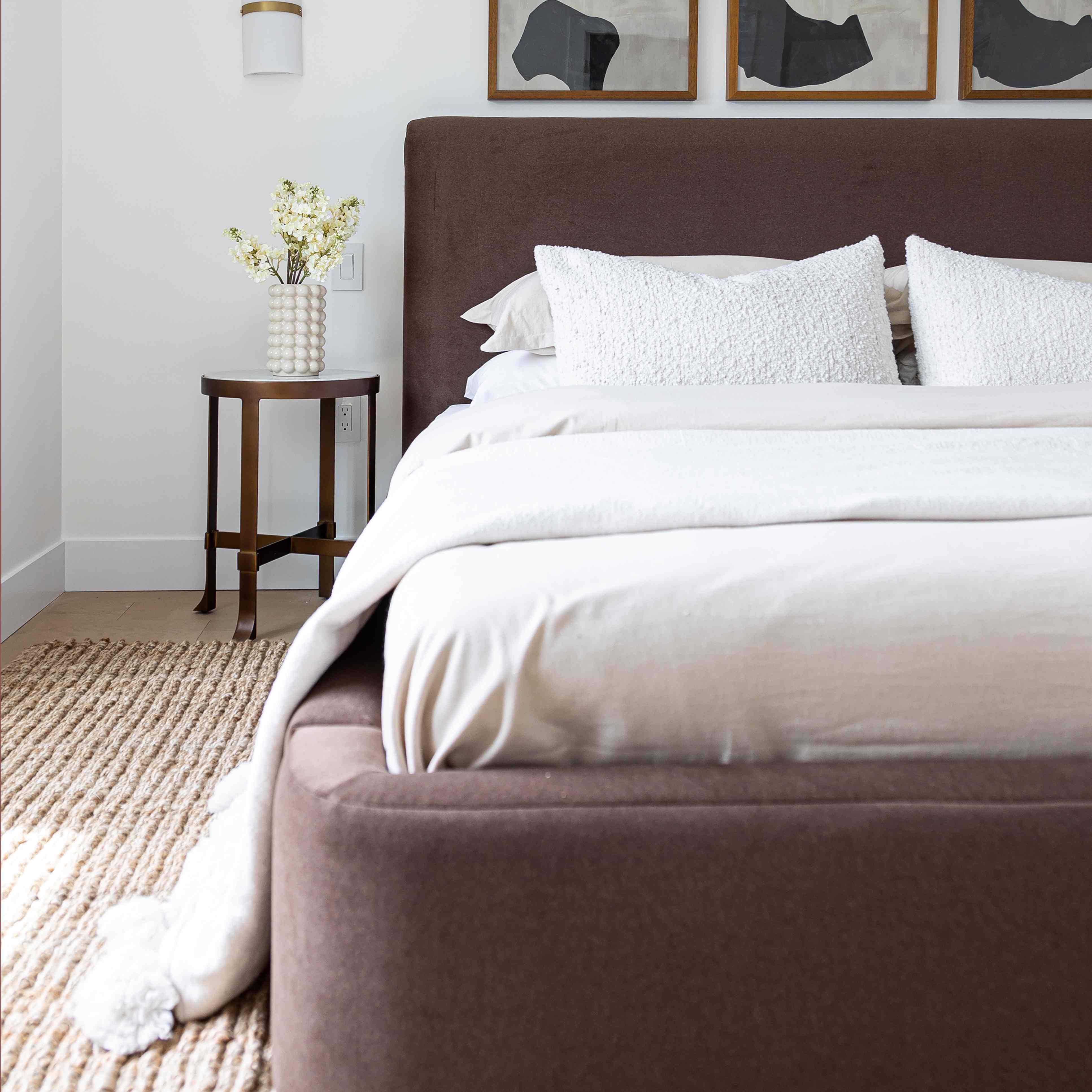 A modern bed with upholstered headboard and footboard in a warm coffee velvet fabric, dressed in white bedding and pillows, set in a bright bedroom with a side table and wall art.