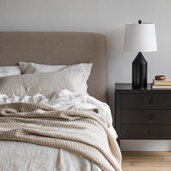 Bedroom with a Lunelle bed featuring beige bedding and pillows, a nightstand with a lamp and books.