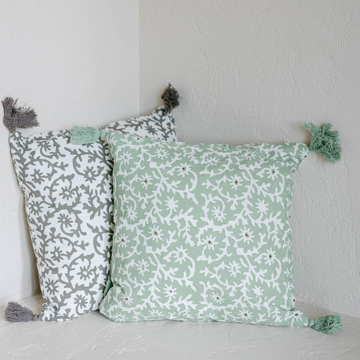 Two patterned pillows with tassels on a light gray surface against a white wall.