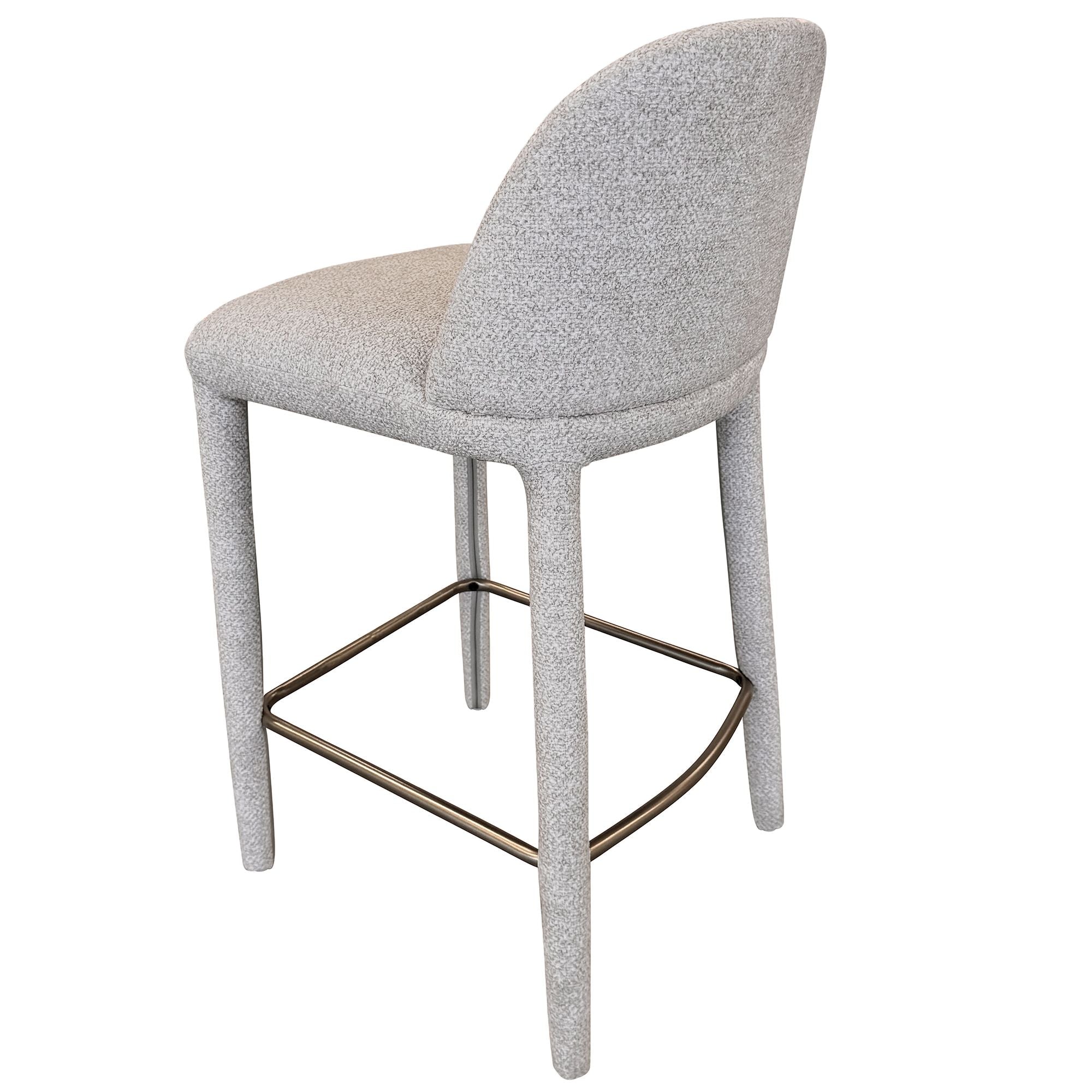 Angled back view of the beige Myla counter stool showing its smooth, continuous upholstered silhouette and brass footrest.