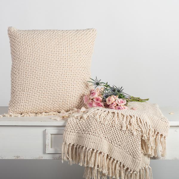 Beige knitted pillow and throw blanket on a bench with pink flowers against a light gray background