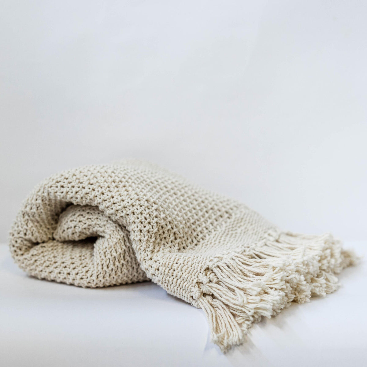 Folded beige knitted blanket with tassels on a light gray background
