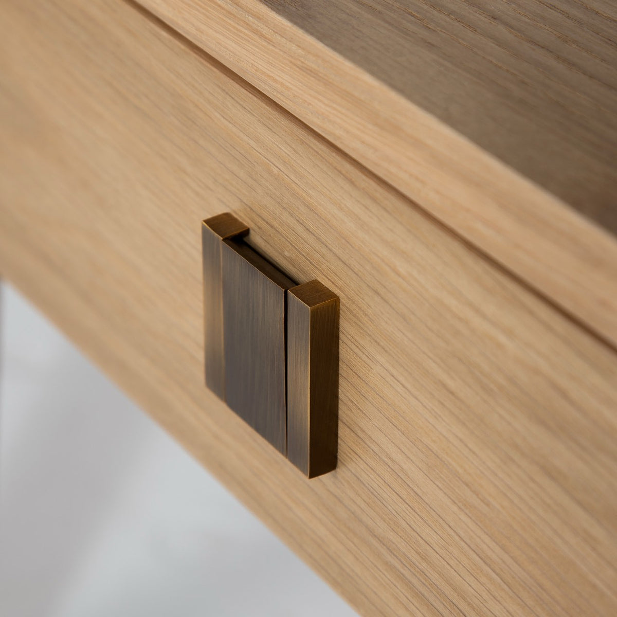 Close-up detail of the Sutton nightstand's square brushed-bronze hardware pull on the natural oak drawer front.
