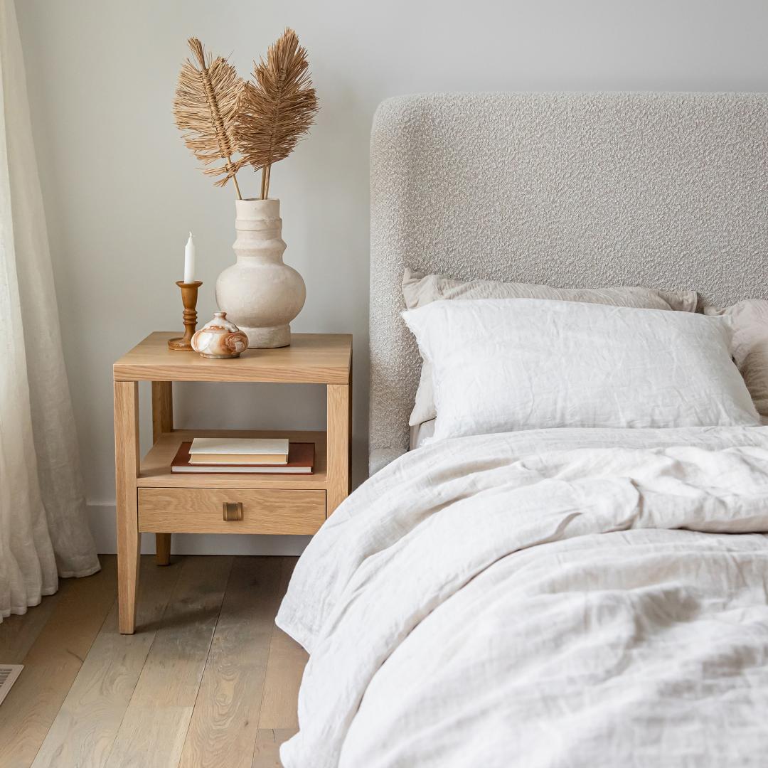 The Sutton oak nightstand styled next to a cream bouclé upholstered bed with linen bedding and natural pampas grass.