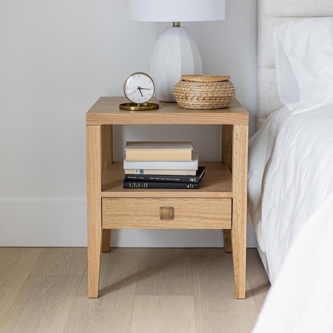 The Sutton oak nightstand styled in a bright bedroom with a white lamp, books, and a woven basket next to a light headboard.