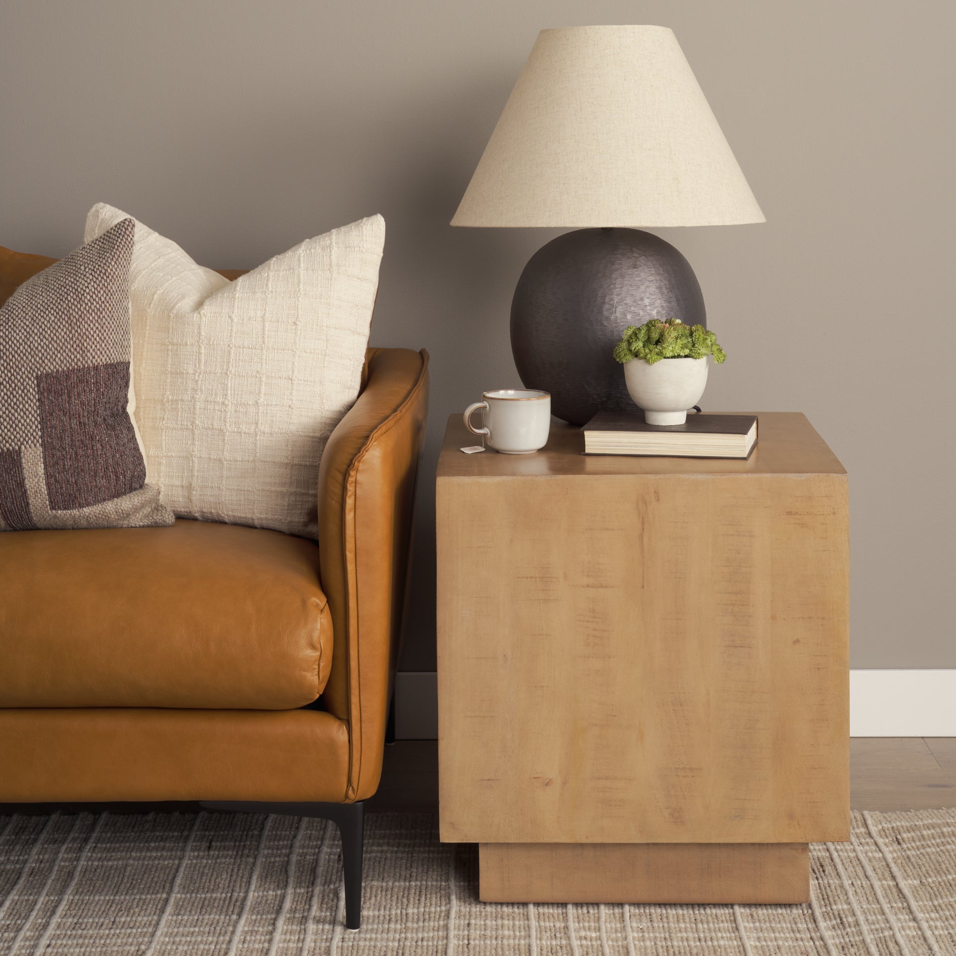 Brown leather sofa with decorative pillows and a wooden side table with lamp, mug, and plant.
