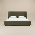Aveline bed with a tall curved headboard and matching footboard in a muted olive green color velvet fabric, set against a plain beige studio background.