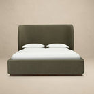 Aveline bed with a tall curved headboard and matching footboard in a muted olive green color velvet fabric, set against a plain beige studio background.