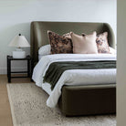 Aveline Velvet bed with a tall curved headboard and matching footboard in dark olive green, styled with white bedding, floral accent pillows, and a dark green throw blanket, set in a bedroom with a nightstand and lamp.