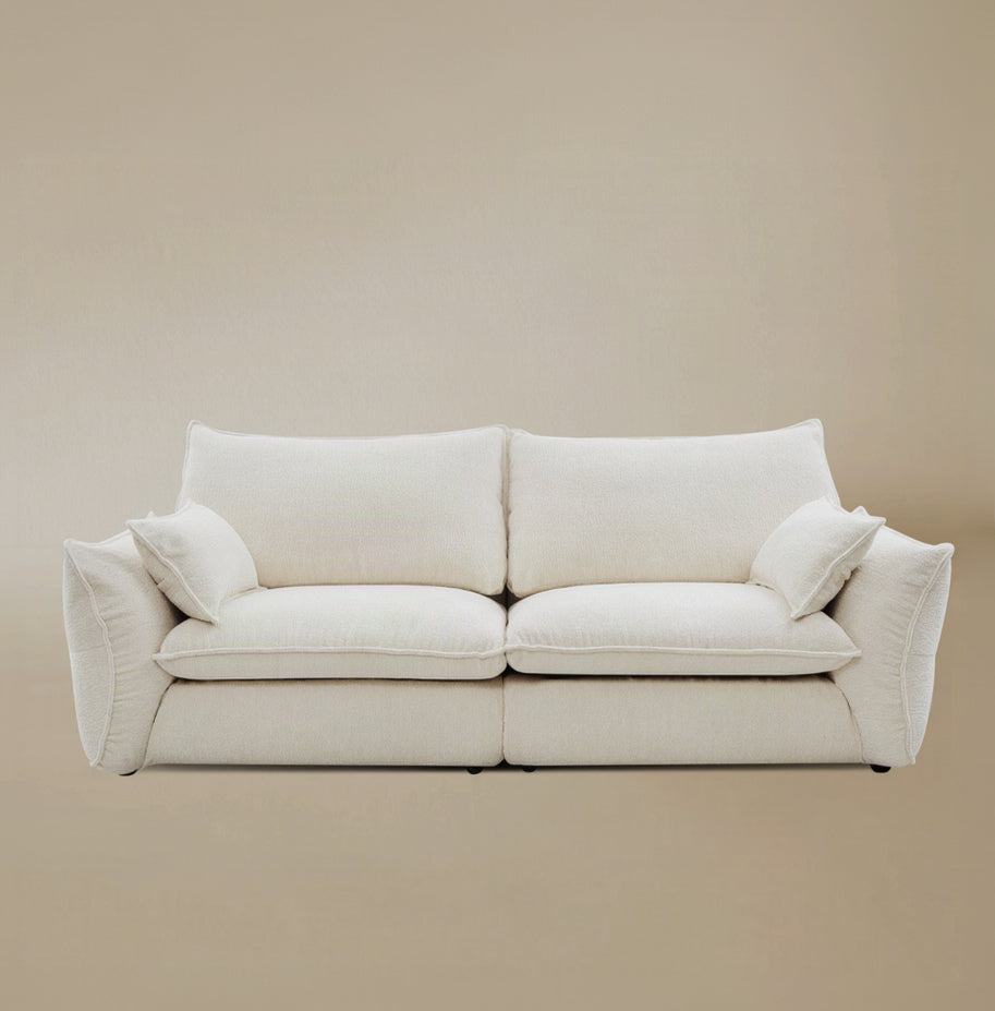 Bella Boucle Sofa upholstered in cream boucle fabric, featuring oversized square back cushions, with matching throw pillows on the side, and flared armrests, set against a solid beige studio background.