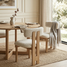White bouclé and natural ash wood dining chairs styled around a matching light wood round dining table in a bright, airy dining room next to a large window...