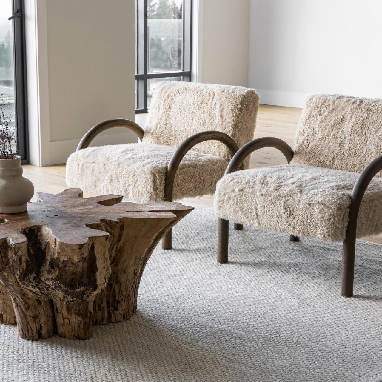 A pair of Coco faux fur chairs styled in a bright, modern living room with a natural wood coffee table and large abstract art.