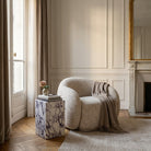 Beige Estelle armchair with a blanket in a stylish room with a Bulgari Charm Side Table and large window.