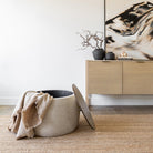 The Haven Storage Ottoman with the wood top removed to show blankets stored inside, styled in a bright minimalist room.
