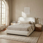 A modern upholstered bed in a warm neutral bedroom setting, featuring a high curved headboard and matching footboard in off-white bouclé fabric, dressed in beige linens and pillows, flanked by wooden nightstands.