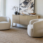 A pair of buff Milo swivel chairs styled in a bright, modern living room with a neutral jute rug and fluted wood sideboard.