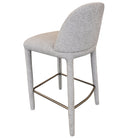 Angled back view of the beige Myla counter stool showing its smooth, continuous upholstered silhouette and brass footrest.