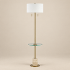 Perrin travertine shelf floor lamp featuring a plated antique brass frame, clear glass shelf, and off-white linen shade on a beige studio background.
