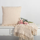 Beige knitted pillow and throw blanket on a bench with pink flowers against a light gray background