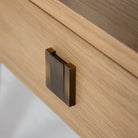 Close-up detail of the Sutton nightstand's square brushed-bronze hardware pull on the natural oak drawer front.