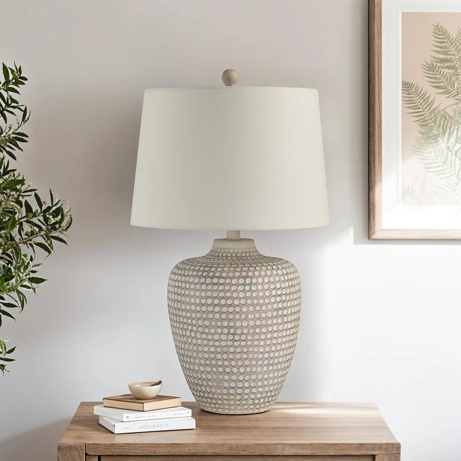 Textured table lamp on a wooden surface with a plant and framed artwork in the background.