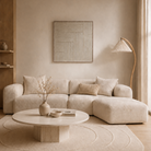 A modern beige sectional sofa in a minimalist, warm-toned living room with a round wooden coffee table, textured beige rug, and a curved floor lamp.